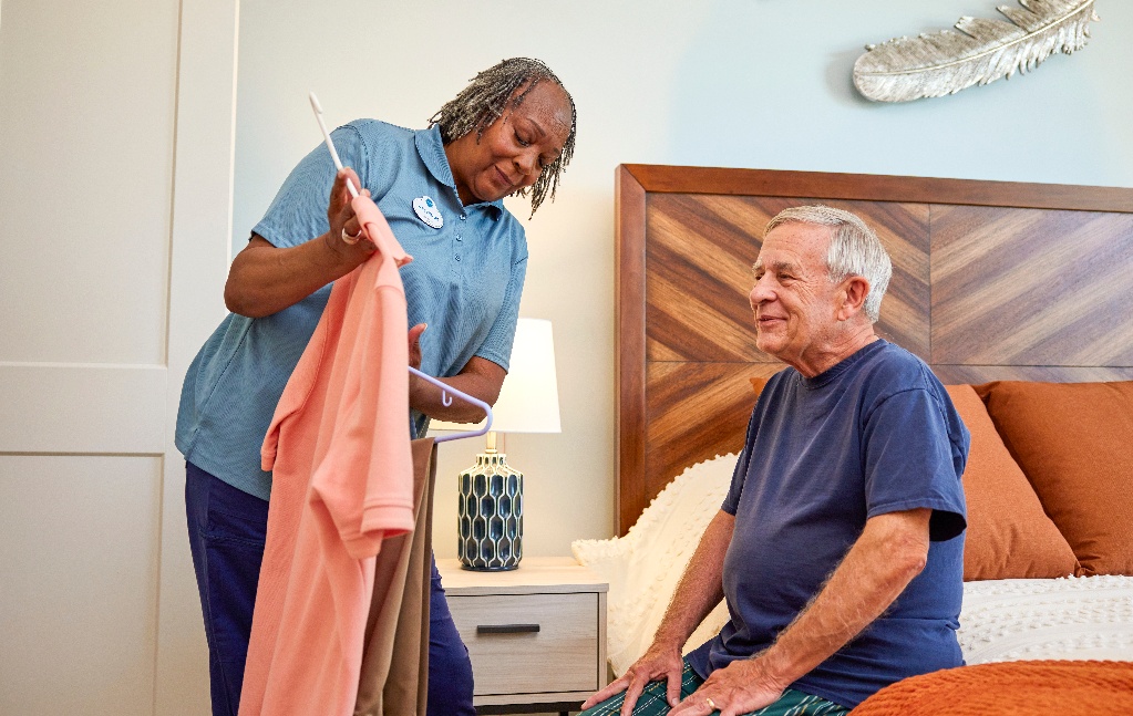 An Arbor caregiver is holding up clothing options for a senior resident sitting on his bed